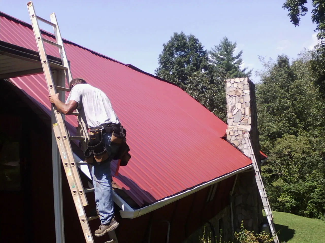 Roofer on a ladder beside a red metal roof and stone chimney — Soffit & Fascia Repair by Roofing Brothers in Allyn